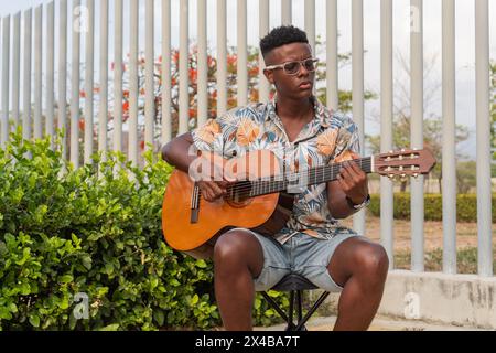A focused young man wearing a tropical shirt strums an acoustic guitar in a serene outdoor setting with lush greenery and modern architecture. Stock Photo
