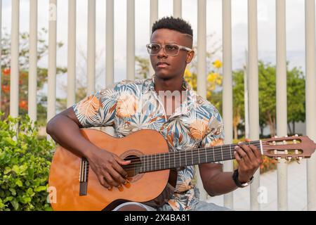 A young African American male musician plays his guitar, seated outdoors with a serene expression and surrounded by a lush urban environment. Stock Photo