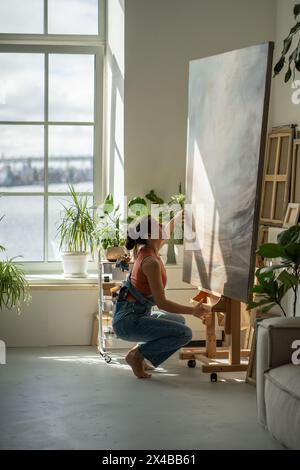 Interested woman artist looking at details oil picture adjusts canvas on easel to continue painting. Stock Photo