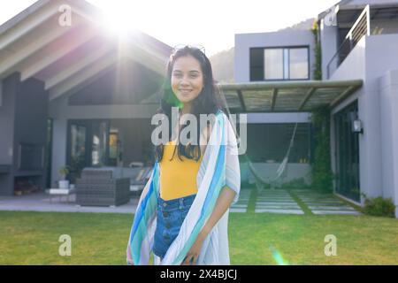 Biracial young woman standing outdoors, smiling in front of house. Wearing a yellow top and denim, she has long dark hair, enjoying sunny day, unalter Stock Photo