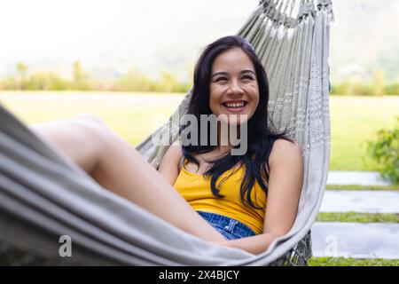 Biracial young woman relaxing outdoors in hammock, looking away with joy. Wearing a casual yellow top and denim shorts, enjoying a peaceful moment, un Stock Photo