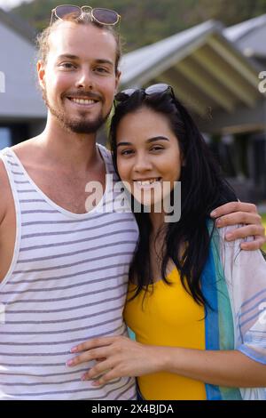 Diverse couple embracing outdoors by pool, smiling at camera. He, light-skinned with beard, wearing a tank top; she, darker-skinned with long dark hai Stock Photo