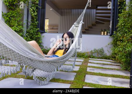 Biracial young woman relaxing outdoors in hammock, using smartphone. Wearing sunglasses and casual clothes, she enjoys a sunny day with long black hai Stock Photo