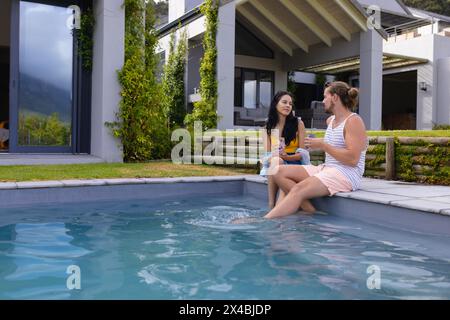Biracial woman and Caucasian man outdoors, sitting by pool, holding drinks, talking, copy space. She has dark hair, he's in pink shorts, both enjoying Stock Photo