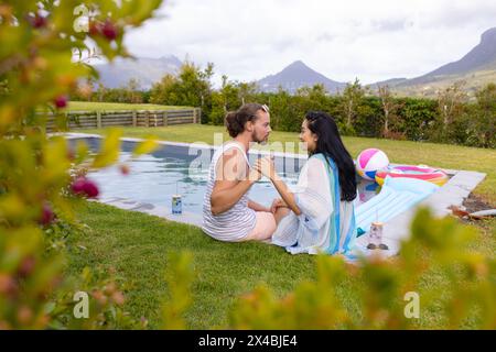 Diverse couple holding hands outdoors by pool, gazing into each other's eyes. She, with long black hair, and he, sporting beard and short hair, enjoyi Stock Photo