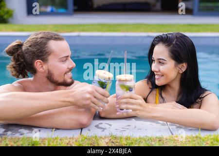 Caucasian male and biracial female enjoying drinks outdoors by pool. Both sporting dark hair, woman with bun and man with ponytail, laughing together Stock Photo