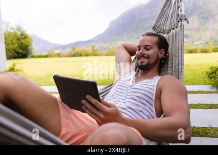 Bearded Caucasian man relaxes in a hammock with a tablet. Wearing casual summer clothes, he enjoying a peaceful moment in a serene blue background, un Stock Photo
