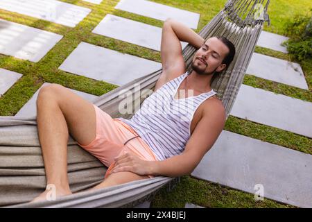Caucasian young man relaxing outdoors in hammock. Wearing casual summer clothes, sporting short brown hair and light stubble, enjoying a peaceful mome Stock Photo