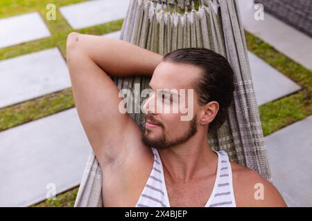 A young Caucasian man relaxing outdoors in hammock, enjoying a swim nearby. Sporting short brown hair and a beard, wearing a white striped tank top, e Stock Photo
