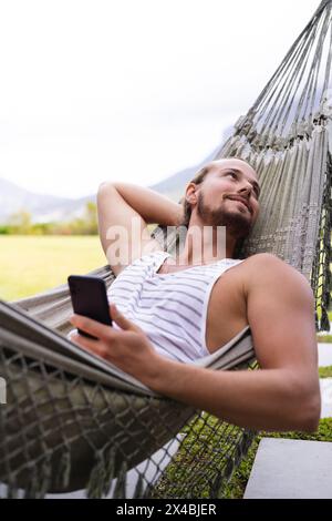 A young Caucasian man relaxing outdoors in hammock, holding a smartphone. Sporting light stubble and long hair, wearing a tank top, enjoying peaceful Stock Photo