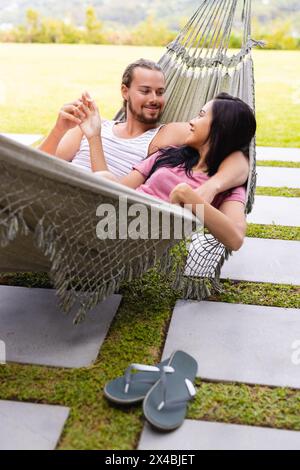 Diverse couple relaxing outdoors in hammock, holding hands. She, with long black hair, and he, sporting shoulder-length brown hair, are enjoying a pea Stock Photo