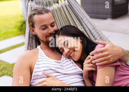 Diverse couple embracing outdoors in hammock, both in casual attire. She, with dark hair, and he, sporting a beard, relaxing together, unaltered Stock Photo