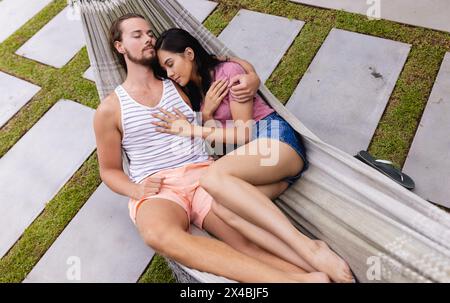Biracial woman and Caucasian man relaxing outdoors on hammock. Both dressed in summer attire, sharing smiles and gentle conversation, surrounded by a Stock Photo