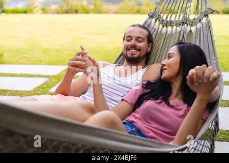 Diverse couple relaxing outdoors in hammock, man holding smartphone. Both with dark hair, she with light brown skin and he with fair skin, enjoying qu Stock Photo