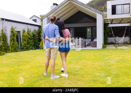 Diverse couple embracing outdoors, enjoying new home view. She, with black hair, and he in a casual blue shirt, sharing a moment in a serene blue back Stock Photo