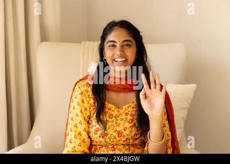 Indian woman in yellow dress waves at camera during home video call. Sporting long black hair and bright smile, she exuding warmth with her dark eyes, Stock Photo