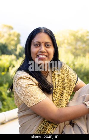 Indian woman wearing traditional attire, smiling outdoors. She has long black hair, bindi, and is wearing a gold and cream saree, unaltered Stock Photo