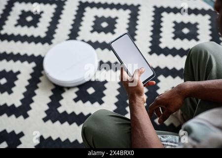 High angle view of unrecognizable Black man holding smartphone using app for robotic vacuum cleaner Stock Photo