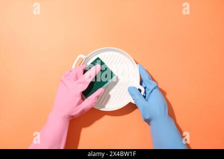 Two hands wearing rubber gloves of two different colors are washing a ceramic plate with a sponge on a red background. Cleaning concept for ads. Stock Photo