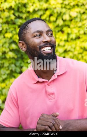 Outdoors, African American man wearing pink shirt, smiling. Sporting short black hair and a beard, he stands before green bushes, exuding warmth, unal Stock Photo