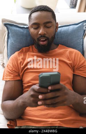 African American homeowner sitting, focused on smartphone. He has short black hair, beard, wearing orange shirt, unaltered Stock Photo