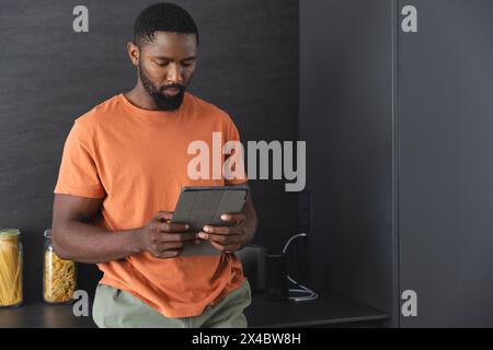 At home, African American male holding tablet in kitchen, copy space. Wearing an orange shirt and sporting short black hair and a beard, he's browsing Stock Photo