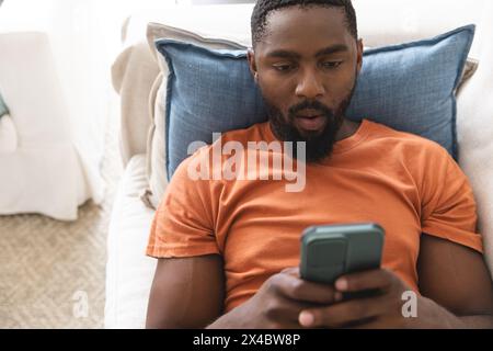 African American male homeowner lying on couch, looking at smartphone. Wearing orange shirt, sporting beard and short black hair, he enjoying quiet mo Stock Photo