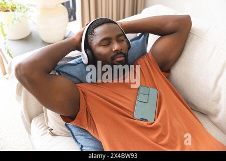 At home, African American male relaxing on couch, wearing headphones. Enjoying music, sporting short black hair and dressed in an orange shirt, he's f Stock Photo