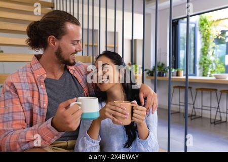 Diverse couple laughing, holding cups, sitting indoors at home. Biracial woman has long black hair, Caucasian man has brown hair and beard, unaltered Stock Photo