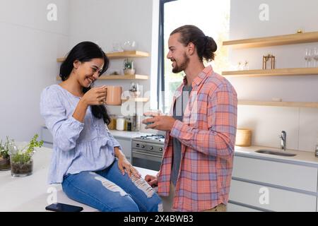 A diverse couple, enjoying coffee together at home. She, with long black hair, and he, sporting beard and long hair, sharing a quiet moment, unaltered Stock Photo