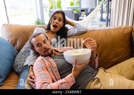 Diverse couple watching TV at home, showing surprise. She has long dark hair, he has beard and long hair, unaltered. Stock Photo