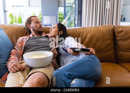 Diverse couple relaxing on couch at home, watching TV and eating popcorn. Biracial woman with long black hair, Caucasian man with beard and curly hair Stock Photo
