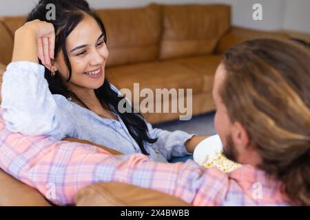 Diverse couple relaxing on couch at home, sharing popcorn. Enjoying cozy moment, both wearing casual clothes, smiling with long black and brown hair, Stock Photo