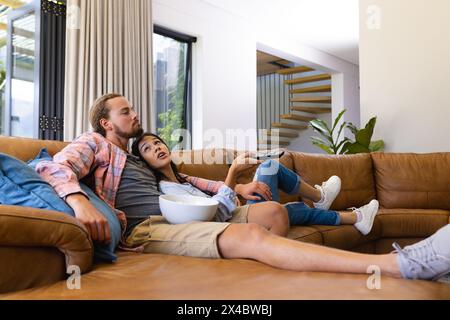 Diverse couple relaxing on sofa at home, watching TV together. She has dark hair, he sports beard, both dressed casually, unaltered Stock Photo