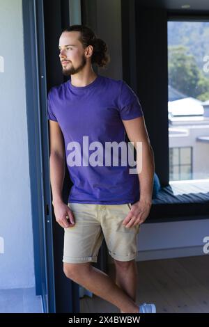 Caucasian young man standing by open door, looking outside at home. He has curly brown hair, beard, and is wearing a purple shirt and beige shorts, un Stock Photo