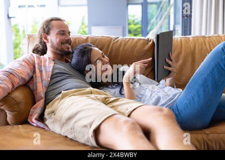 Diverse couple relaxing on sofa at home, looking at tablet. She has dark hair and he sports beard and long hair, unaltered Stock Photo