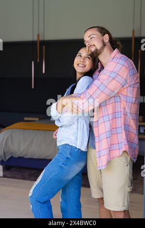 Caucasian male and biracial female couple are embracing, smiling at home. Both have dark hair, he has a beard, she wears minimal makeup, unaltered. Stock Photo