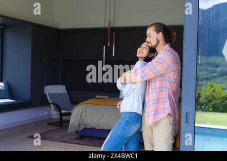 Diverse couple embracing in modern bedroom at home, copy space. She has curly brown hair, he has a beard and long hair, unaltered Stock Photo