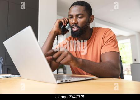 African American male homeowner talking on phone, using laptop at home. He has beard, wearing orange shirt, focused on screen, unaltered Stock Photo
