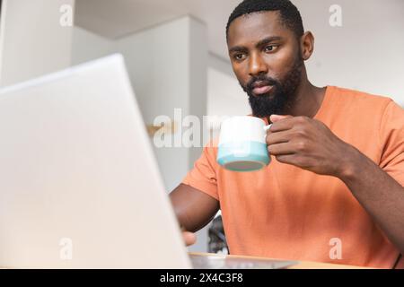 At home, African American male holding cup, looking at a laptop. Wearing an orange shirt and sporting a beard, focusing intently on screen with an ora Stock Photo