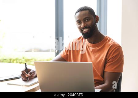 At home, African American male homeowner working on laptop at a desk. Wearing an orange shirt and sporting a beard, he's smiling warmly, creating a fr Stock Photo