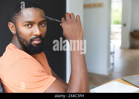 At home, African American male holding pen, looking at camera. Wearing an orange shirt, sporting short black hair and a beard, exuding confidence, una Stock Photo