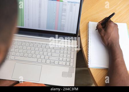 At home, African American male wearing an orange shirt, writing in notebook. Focusing on a spreadsheet, he's also looking at a laptop, deeply absorbed Stock Photo