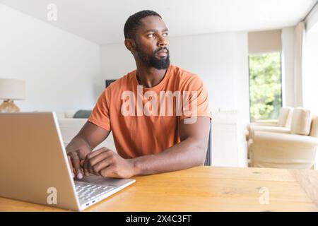 At home, African American male homeowner using laptop at a table. Sporting a beard and short hair, wearing an orange shirt with an orange background, Stock Photo