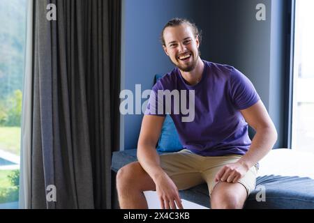 Caucasian young man sitting, smiling, wearing casual clothes at home, copy space. He has a beard, long hair tied back, and is in a well-lit room, unal Stock Photo