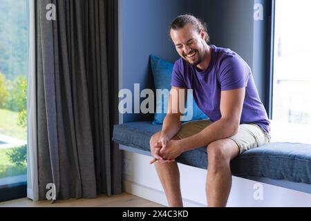 A young Caucasian man, sitting by window, laughing in a light room at home, copy space. Wearing casual clothes, he has short brown hair and a beard, e Stock Photo