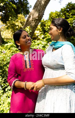 Mother and teenage daughter, both Indian, holding hands, smiling outside. Wearing traditional dresses, mother in pink and teenage daughter in blue, ag Stock Photo