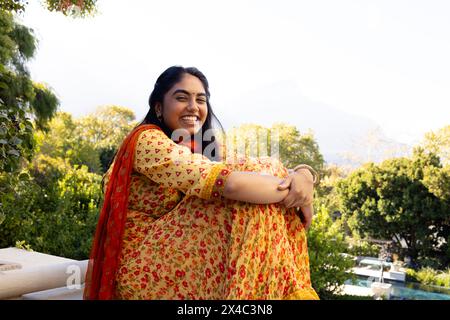A young Indian woman in orange and red laughing outdoors on balcony. Sporting long black hair and a bright smile, she's enjoying sunny weather, unalte Stock Photo