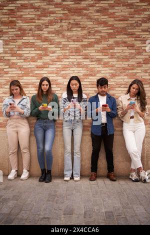 Multiracial serious group young generation z leaning on brick wall using phones outdoors.  Stock Photo