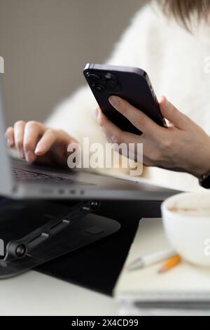 Woman looking worried at cell phone, holding cup of coffee and sitting in front of laptop screen Stock Photo